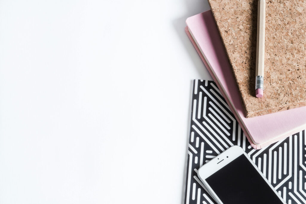 A background image for the introduction of Indigo Design Creative, a local small business that provides design services; a pencil rests on two stacked notebooks beside a black-and-white patterned folder—perfect for a designer—alongside a smartphone on a white surface.